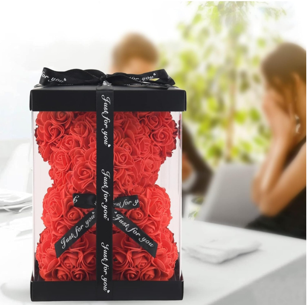 Red rose teddy bear in a clear box with 'Just for you' tag, blurred background of two people sitting.
