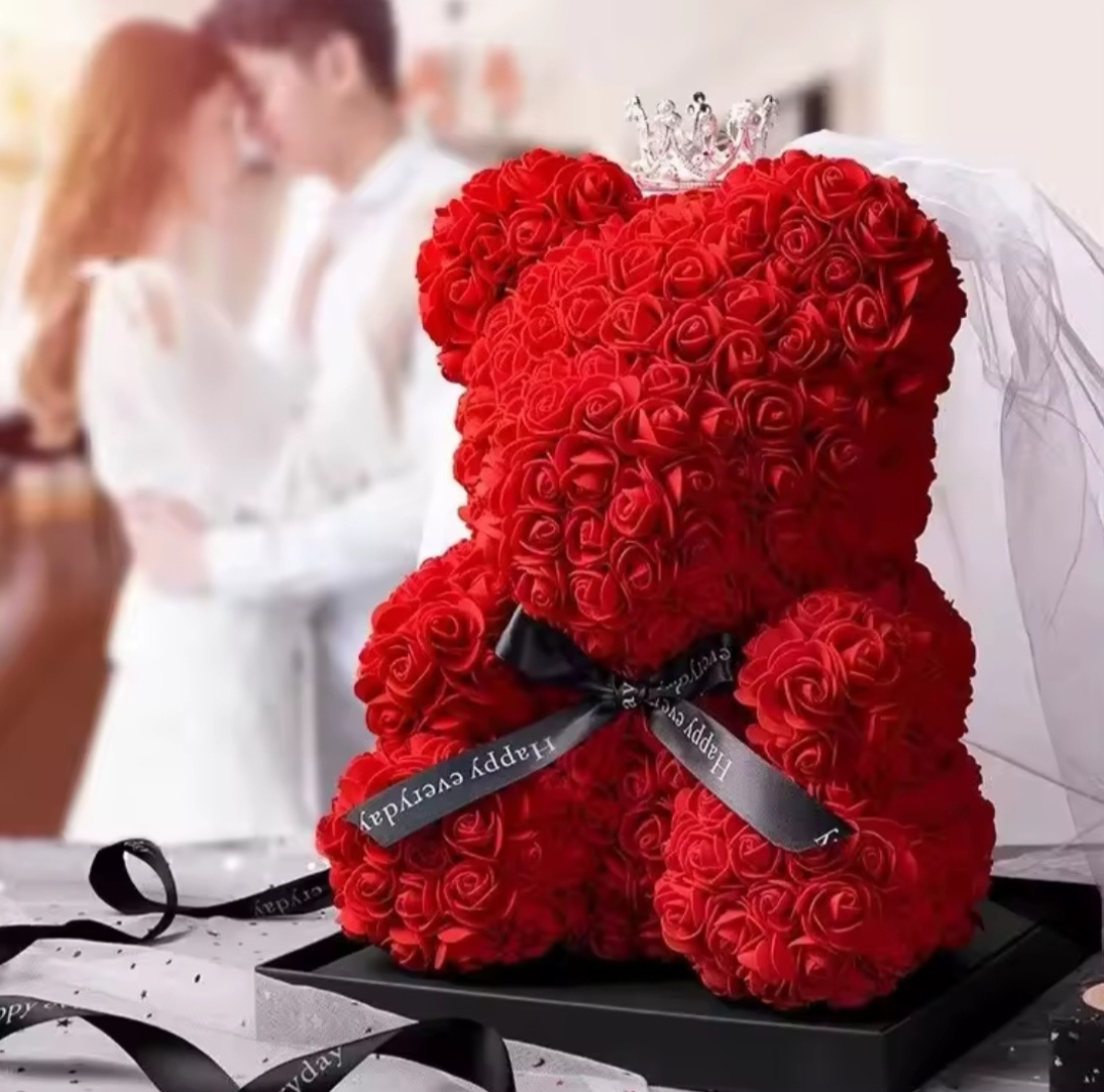 Red rose teddy bear with a tiara and bow, placed on a table with a blurred background of people.