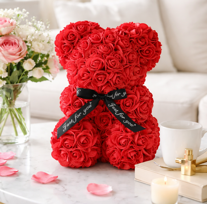 Red rose teddy bear with a black ribbon on a table with flowers and a cup.