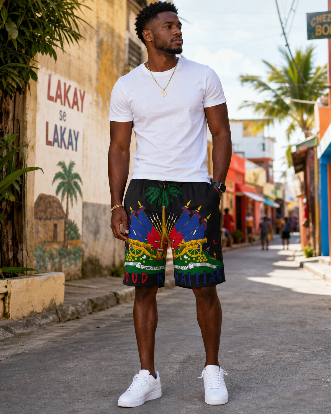 Haitian man wearing black Haiti flag graphic shorts, a plain white T-shirt, and white sneakers while standing on a colorful Caribbean-style street.