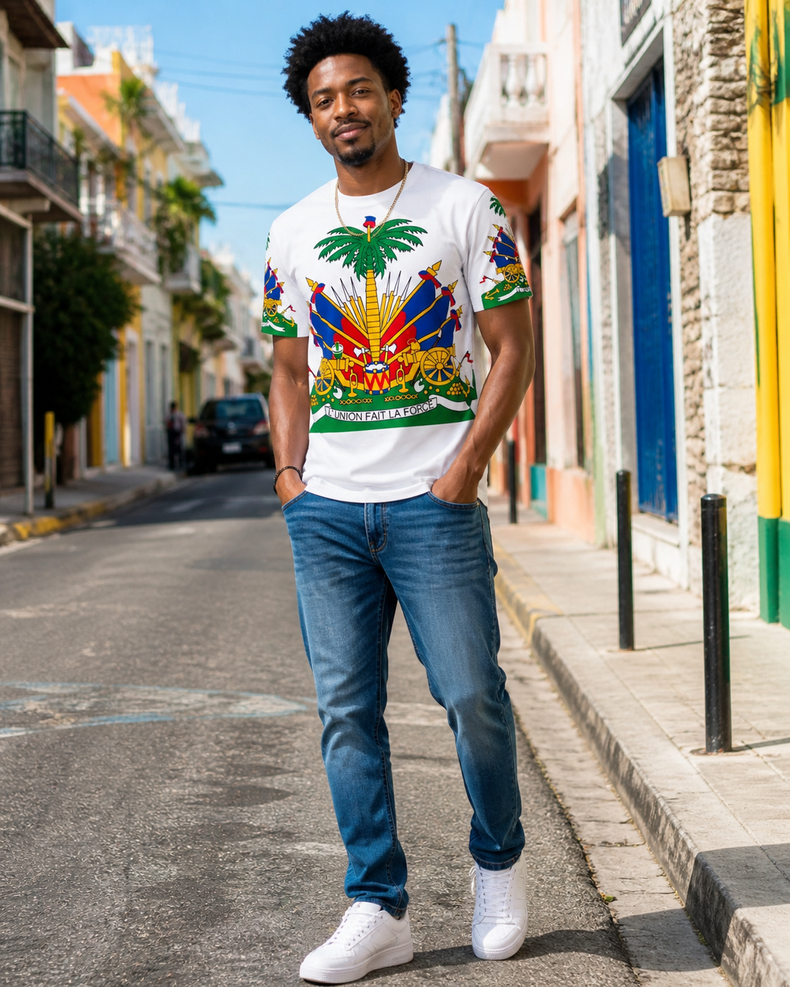 Haitian man with an afro wearing a white Haitian flag graphic T-shirt, blue jeans, and clean white sneakers while standing on a colorful Caribbean-style street.